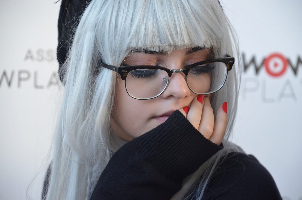 Joven con cabello plateado y gafas, tocándose la boca con una mano.